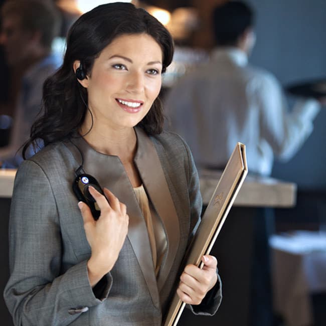 Žena s jemným úsměvem, používající radiostanici Motorola CLP446 se sluchátkem v uchu a drží menu v elegantní restauraci.