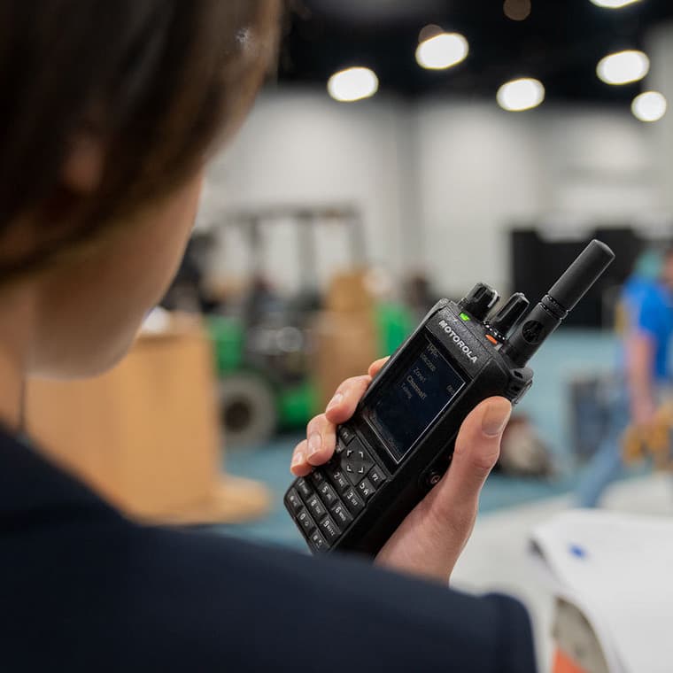 Detailní pohled přes rameno uživatele držícího radiostanici Motorola MOTOTRBO R7 FKP Premium v prostředí haly.