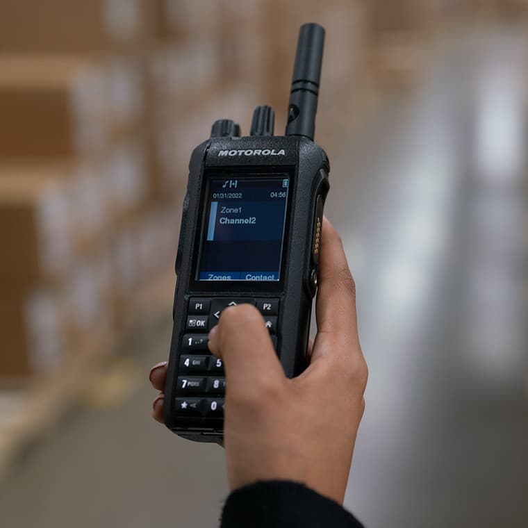 Detail ruky držící radiostanici Motorola MOTOTRBO™ R7 FKP aktivně ovládanou v interiéru skladových prostor.