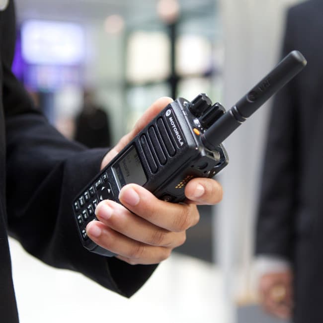 Ruka držící radiostanici Motorola MOTOTRBO DP4801e UHF, záběr z boku s viditelným displejem a klávesnicí, v pozadí postava v obleku.